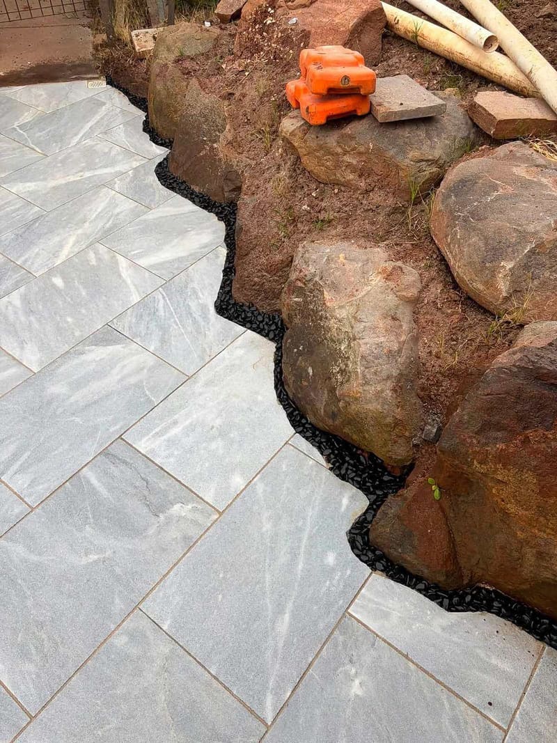 Marble-look pavers scribed around natural boulders with a black pebble border — May Paving